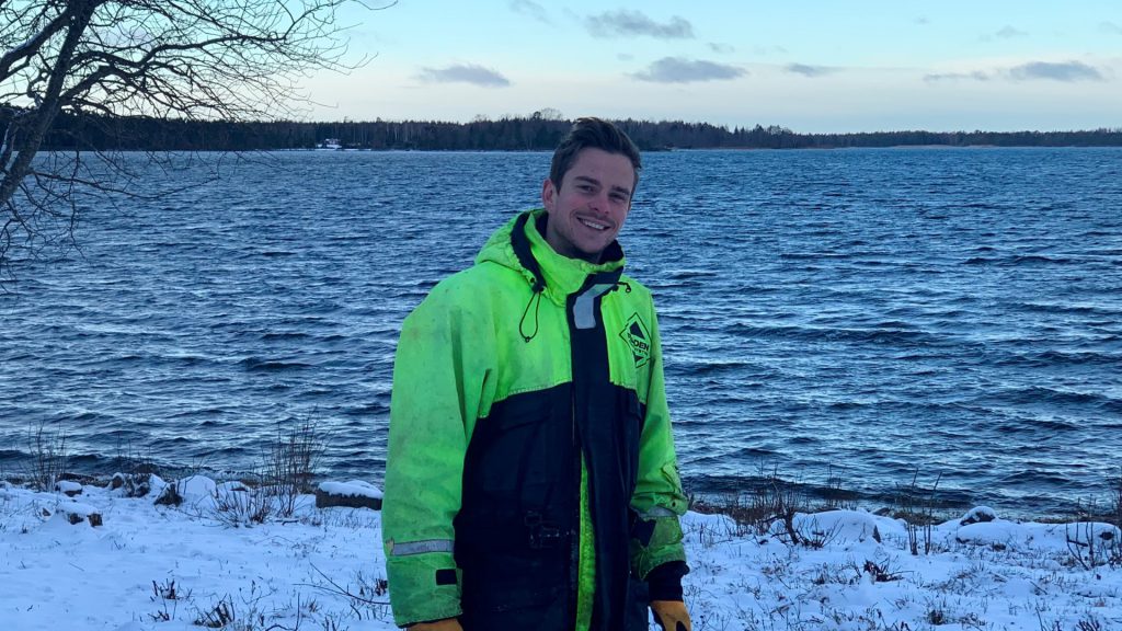 Flexens' Head of Commercial and Energy Modelling Specialist Edvard Nordlund standing by the sea in the Åland Islands, wearing a high-visibility winter jacket on a snowy shoreline.