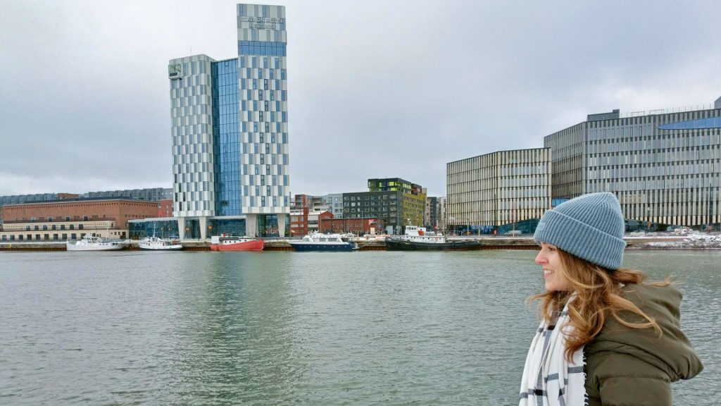 A woman in a gray knit beanie and a green winter coat stands by the waterfront, looking toward the city skyline. Modern office buildings, boats, and an overcast sky are visible in the background.