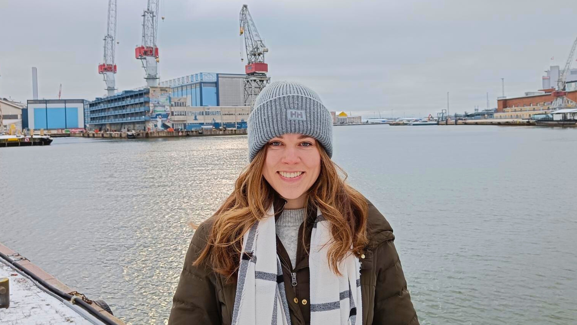 Szilvia, a Project Manager at Flexens, smiles by the Helsinki waterfront, with shipyard cranes and industrial buildings in the background on a winter day.